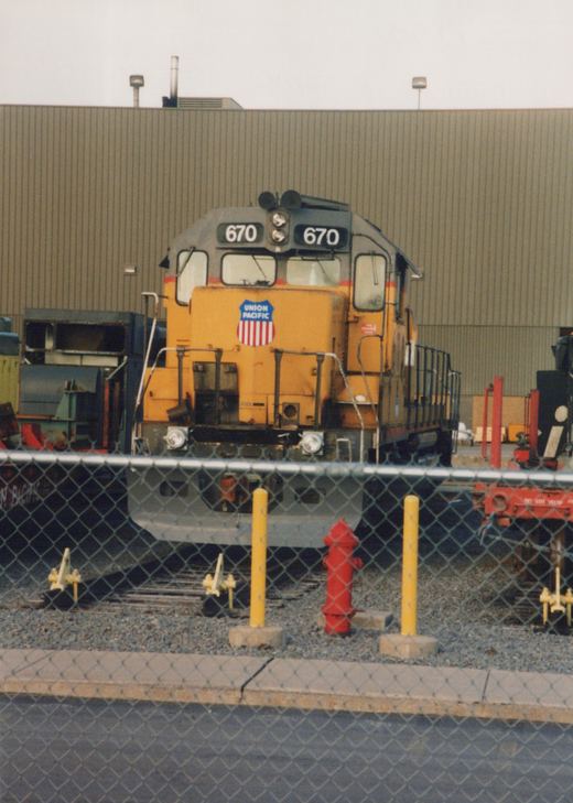 Front view of a yellow and grey Union Pacific diesel locomotive, number 670, parked on tracks behind a chain-link fence with a red fire hydrant and yellow bollards in the foreground.