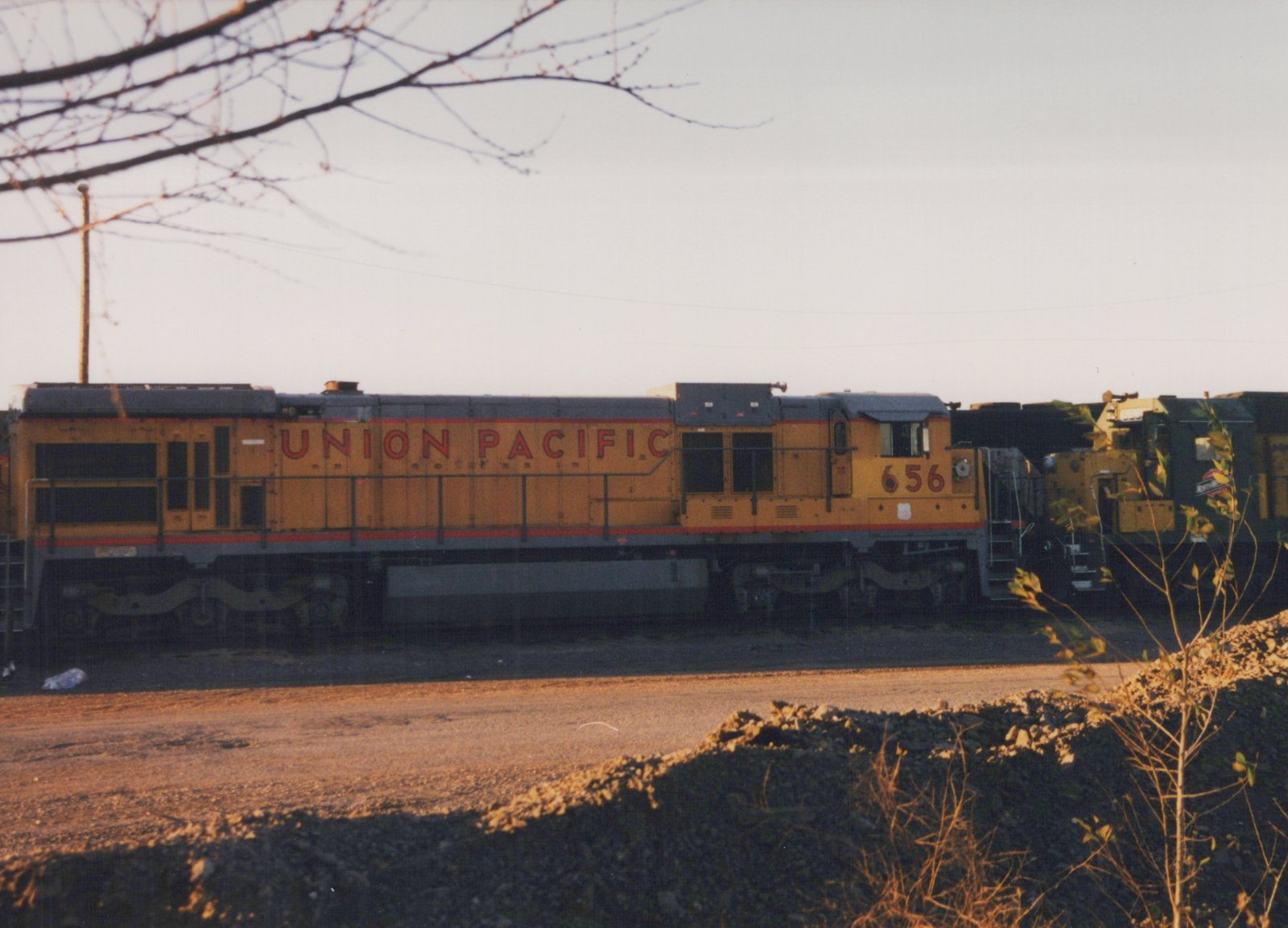 Side view of a yellow Union Pacific diesel locomotive, number 656, parked on a track in a gravel yard with bare trees in the foreground at dusk.