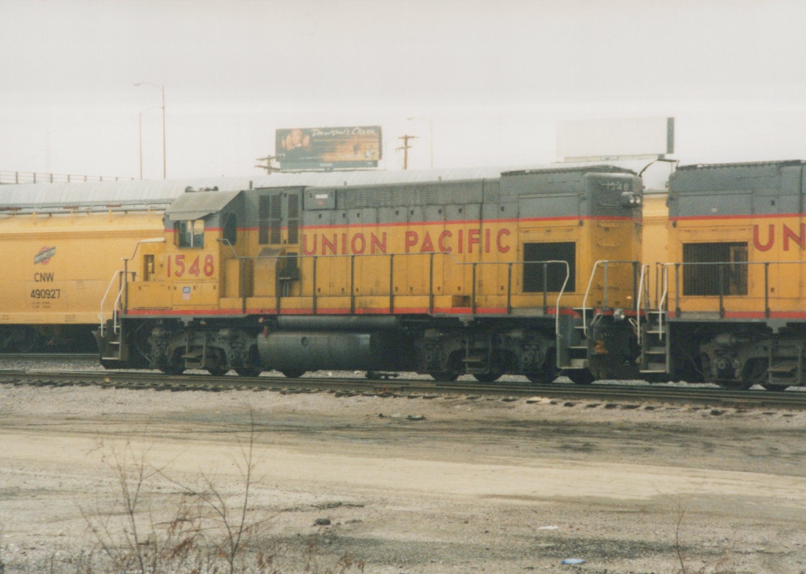 A side view of a yellow and gray Union Pacific diesel locomotive with the number 1548, parked in a gravel yard next to a yellow freight car.