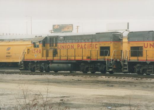 A side view of a yellow and gray Union Pacific diesel locomotive with the number 1548, parked in a gravel yard next to a yellow freight car.