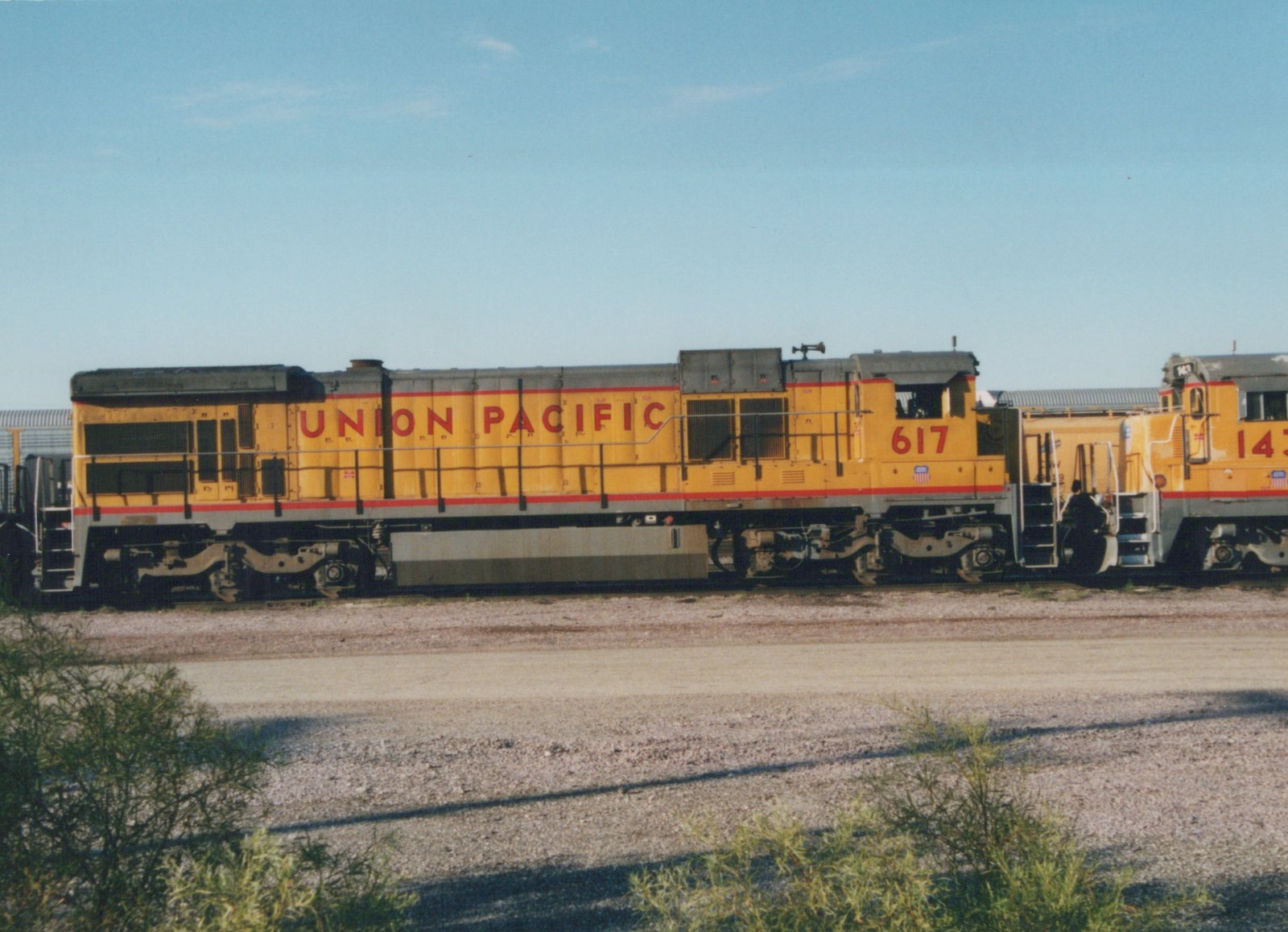 Two yellow Union Pacific diesel locomotives, numbered 617 and 143, are shown side by side in a gravel yard on a sunny day.