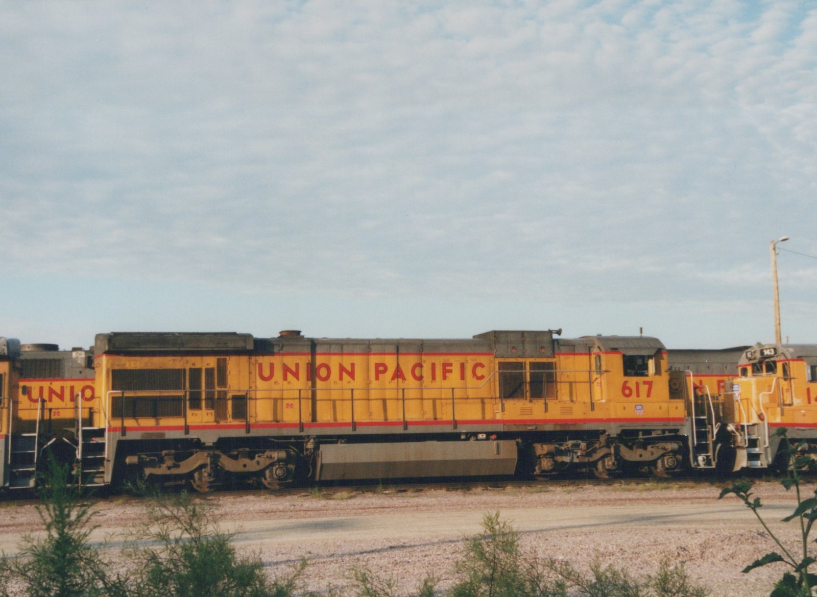Two yellow and gray Union Pacific diesel locomotives, numbers 617 and 143, are parked side by side in a gravel yard under a partly cloudy sky.