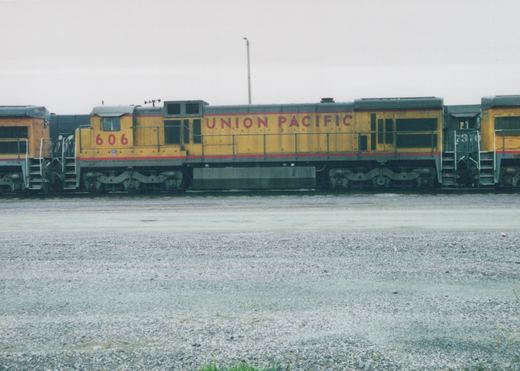 Side view of a yellow and gray Union Pacific diesel locomotive, number 606, parked in a gravel yard with another locomotive behind it, both bearing the 'Union Pacific' name in red lettering.