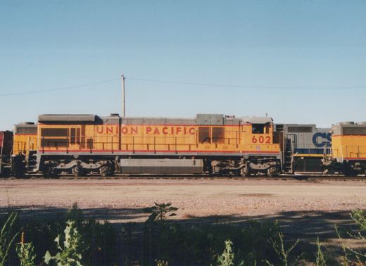 A side view of a yellow Union Pacific diesel locomotive numbered 602, with a gray CSX locomotive visible behind it, set against a clear blue sky in a gravel yard with green weeds in the foreground.