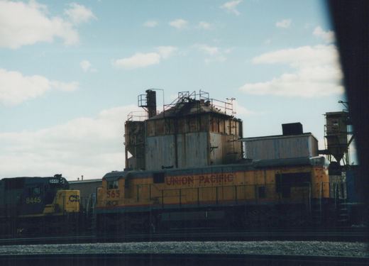Two diesel locomotives, one yellow with 'Union Pacific' lettering and the other dark blue with 'CSX', sit on parallel tracks in an industrial yard under a cloudy sky.