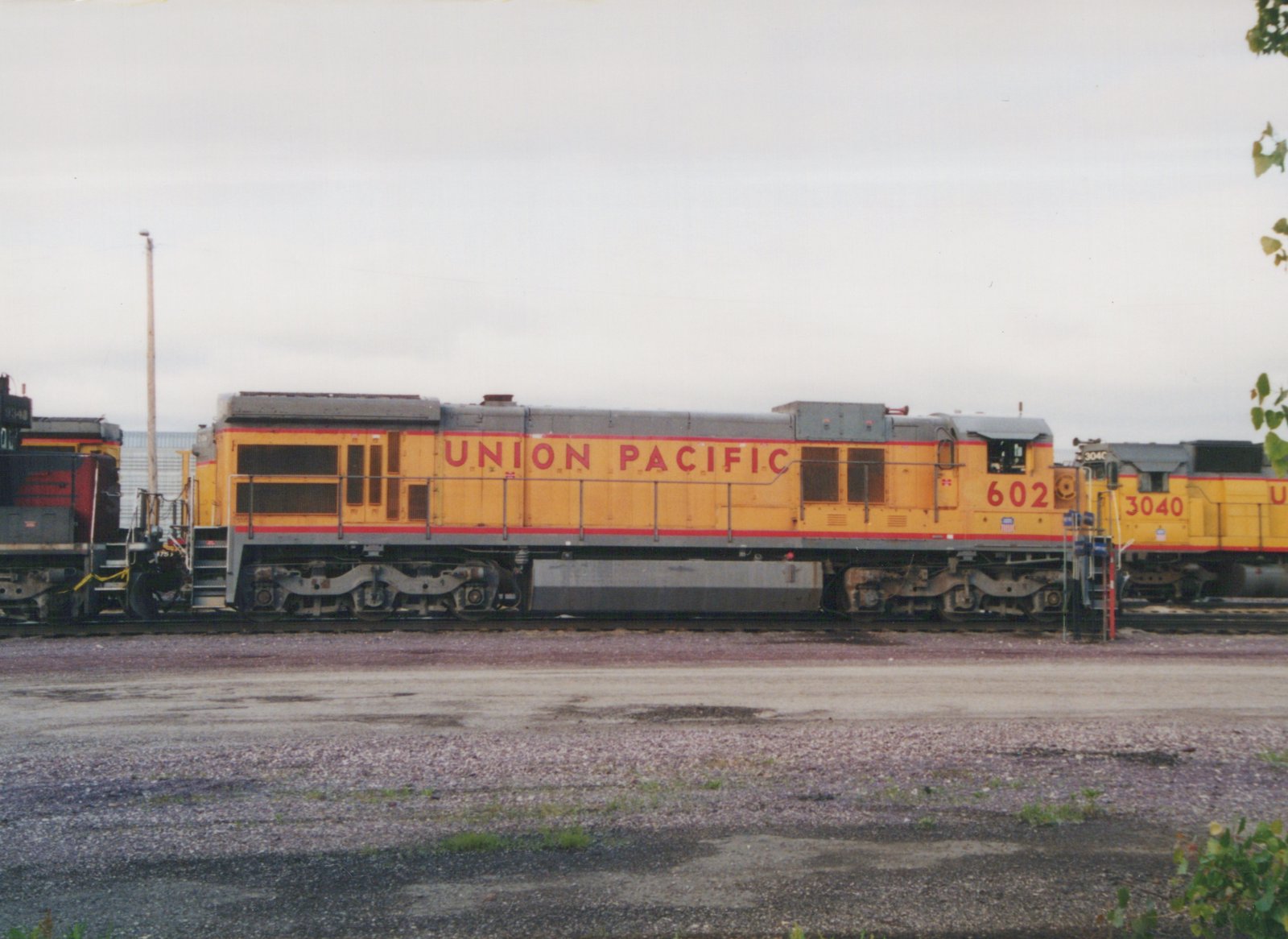 A side view of two yellow, red and gray Union Pacific diesel locomotives, numbered 602 and 3040, standing on parallel tracks in a gravel yard under a gray sky.