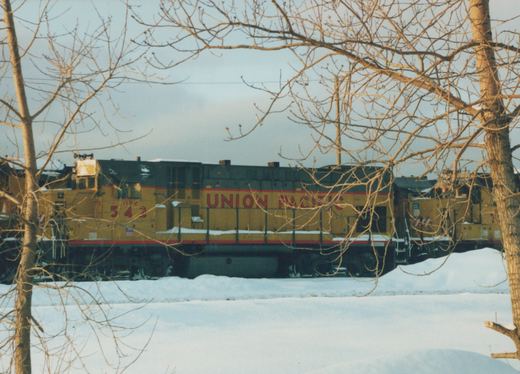 A yellow and red Union Pacific diesel locomotive numbered 542 sits on a track in a snowy landscape, framed by bare tree branches.