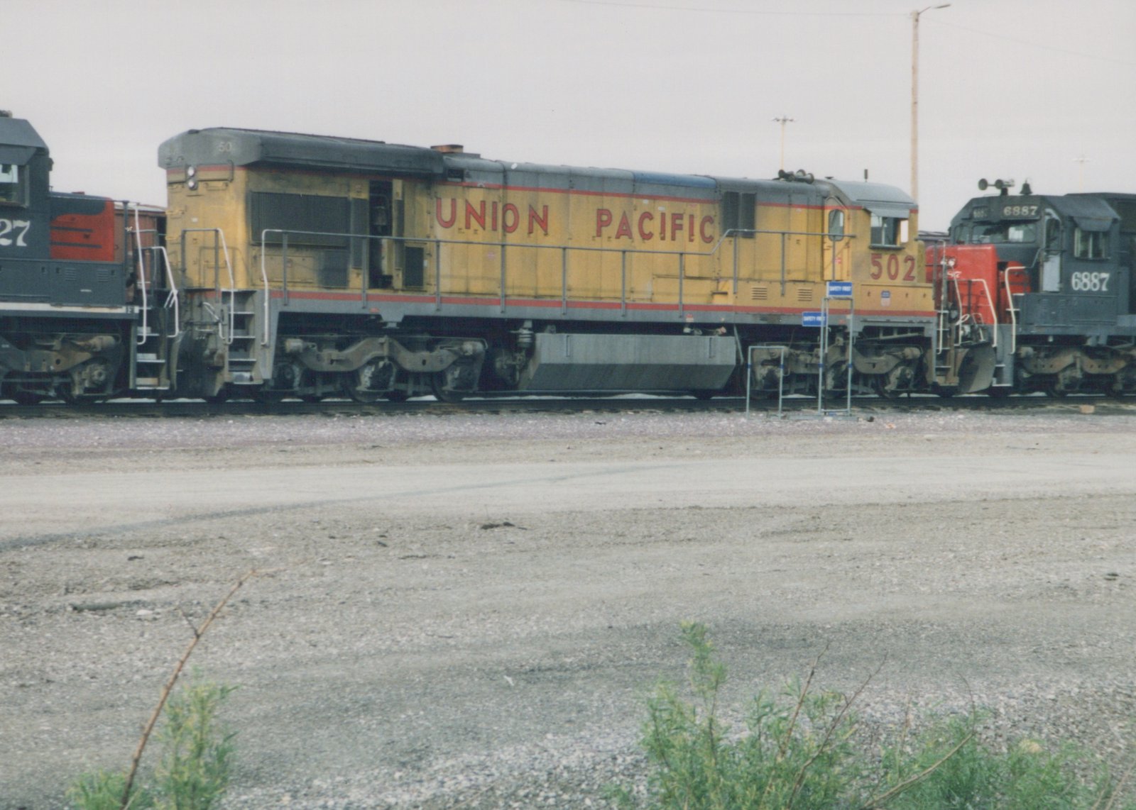 A yellow Union Pacific diesel locomotive, number 502, stands in a gravel yard with other locomotives nearby on a cloudy day.