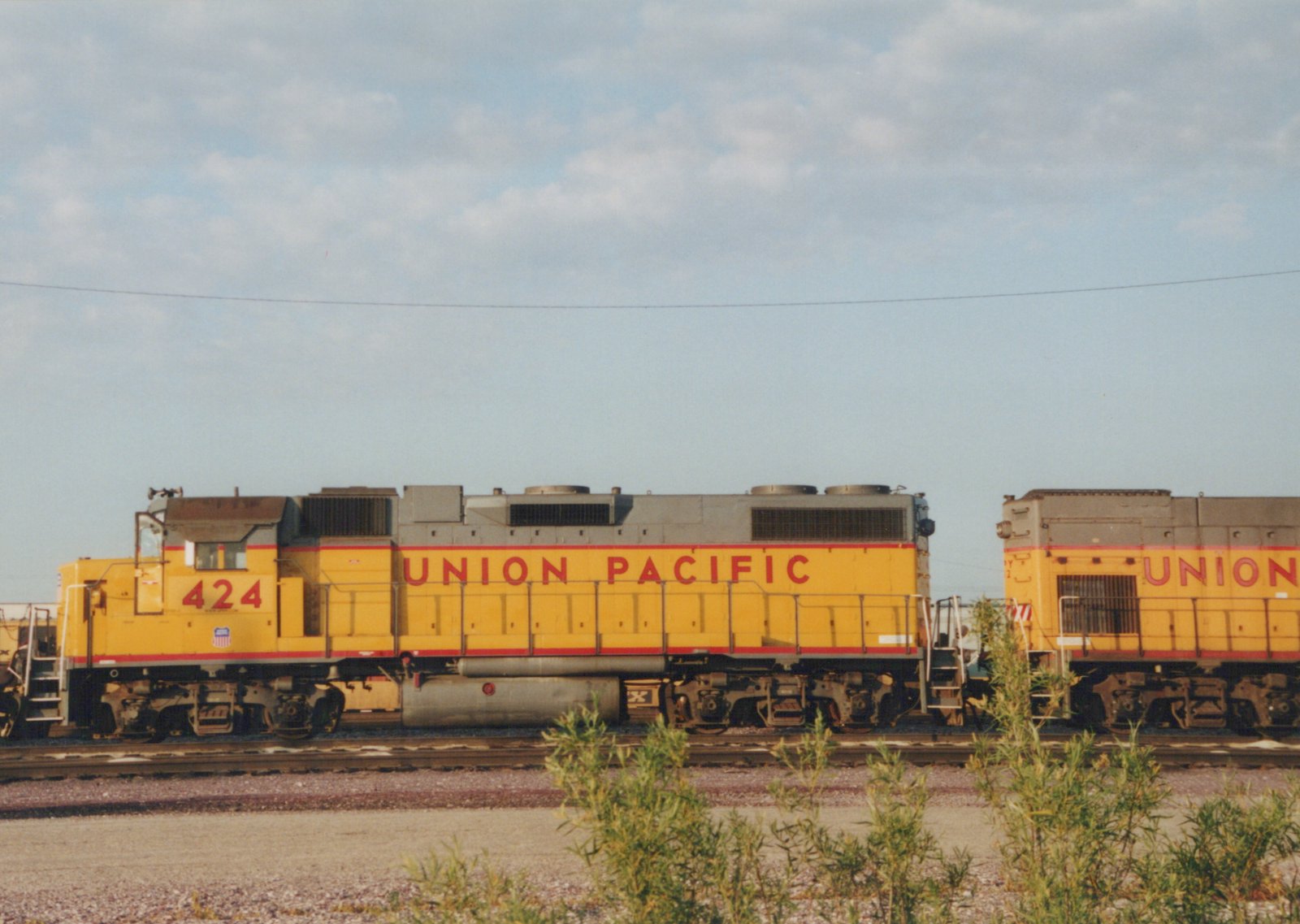 A yellow and red Union Pacific locomotive, number 424, is parked on a track in a rail yard with green shrubs in the foreground and a clear sky.