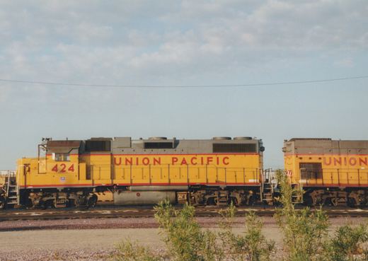 A yellow and red Union Pacific locomotive, number 424, is parked on a track in a rail yard with green shrubs in the foreground and a clear sky.