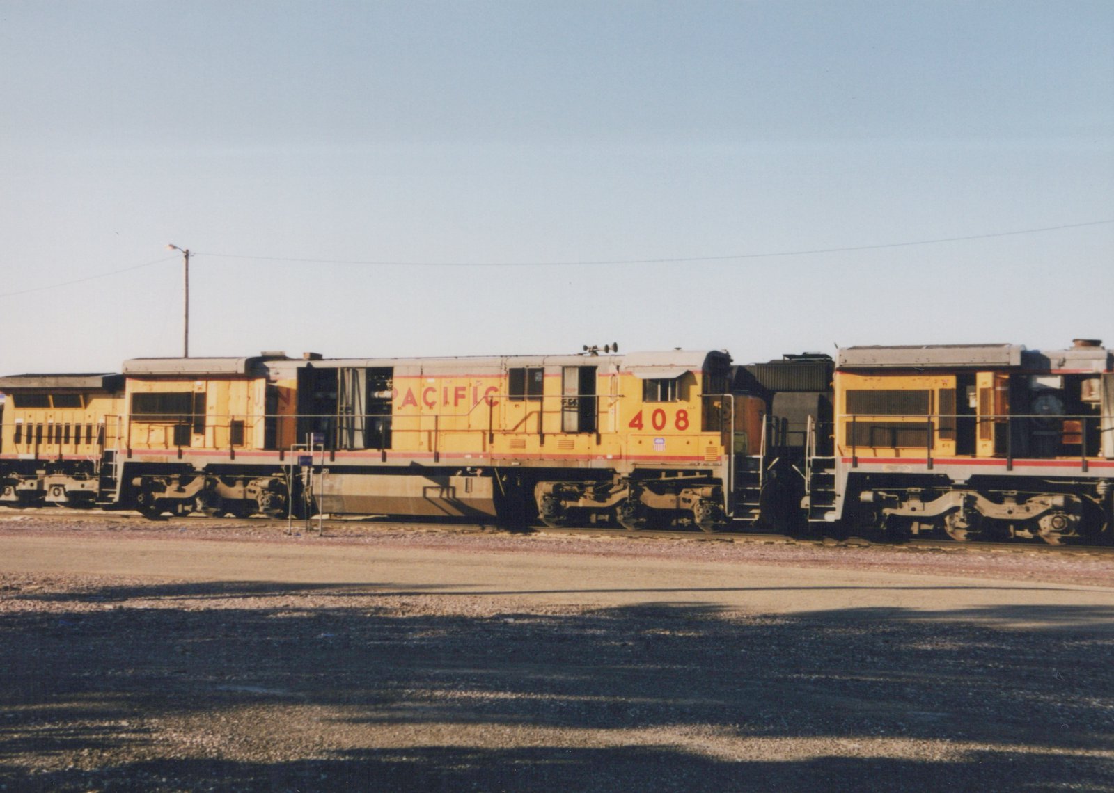 Side view of a yellow Union Pacific diesel locomotive with red stripes and 'PACIFIC' lettering, numbered 408, parked in a gravel yard next to another similar locomotive under a clear sky.