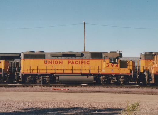 A yellow Union Pacific locomotive with the number 394 on its cab side, sitting on tracks in a gravel yard under a clear sky.