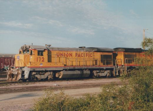 Side view of two yellow Union Pacific locomotives, numbers 172 and 617, standing on tracks in a yard with green bushes in the foreground and a maroon freight car in the background under an overcast sky.