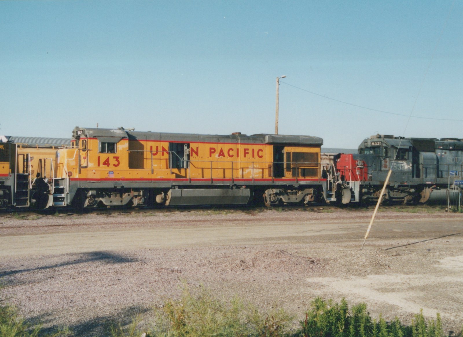 Side view of a yellow Union Pacific locomotive 143 in a rail yard, with a dark gray Southern Railway locomotive 832 behind it on a clear summer day.