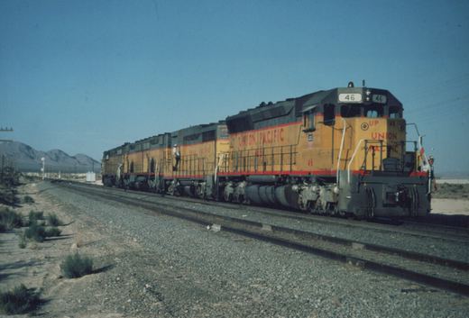 A side view of a yellow and red Union Pacific diesel locomotive, number 46, on a railroad track in a desert landscape with mountains in the background under a clear sky.