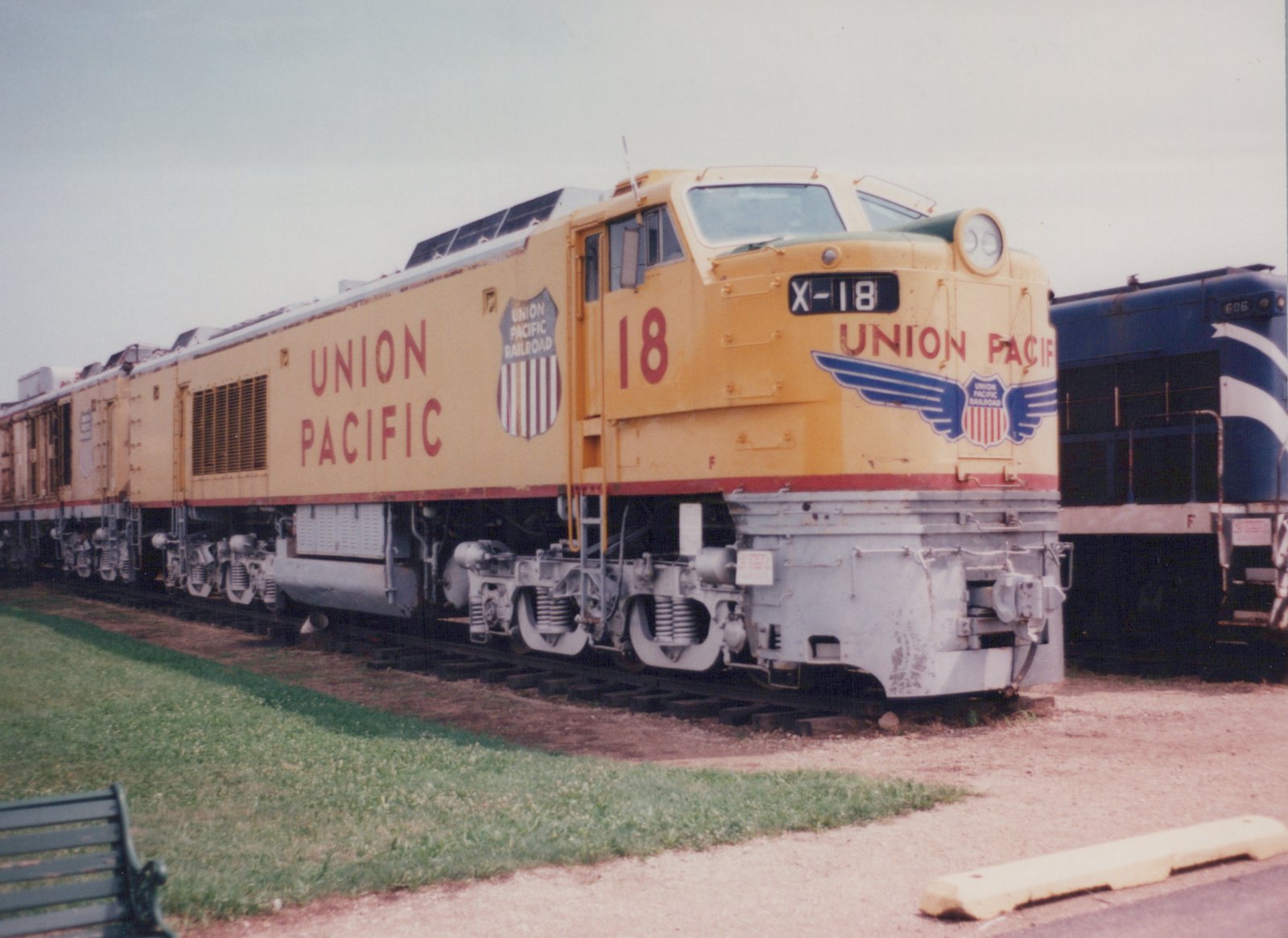 A yellow Union Pacific diesel locomotive, number 18, parked on a track next to a grassy area, with another blue locomotive partially visible in the background.
