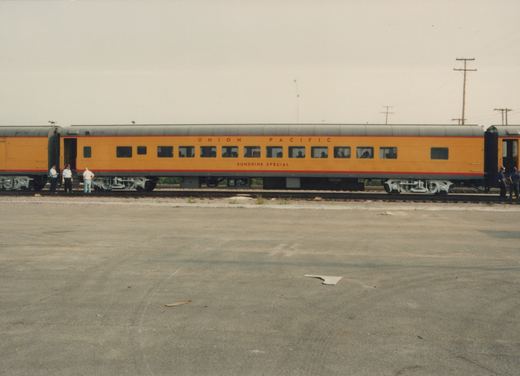 Side view of a long, orange Union Pacific passenger car labeled 'Sunshine Special' with people standing near the open doors, on a gravel yard under an overcast sky.