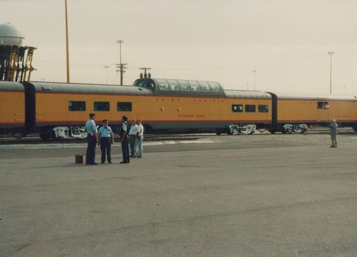 A side view of a Union Pacific passenger train with a distinctive dome car and another passenger car, stopped in a yard with several people around.