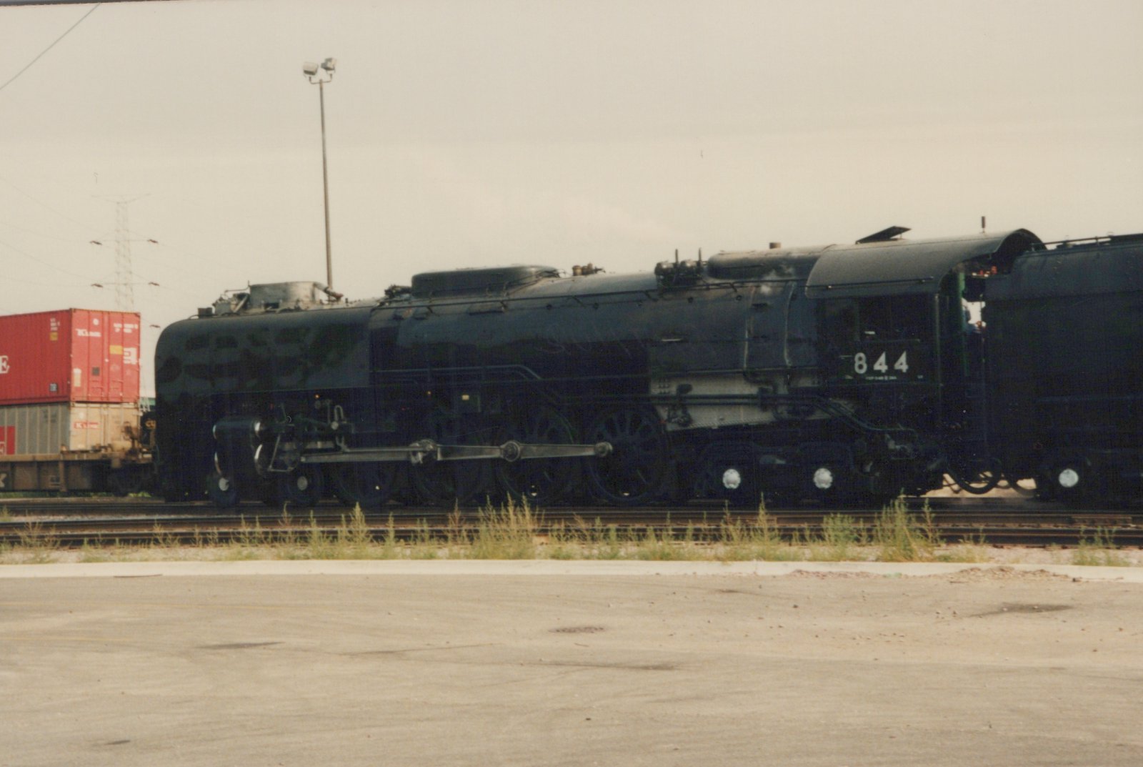 A large black steam locomotive with the number 844 on its cab side is parked on tracks in a rail yard, with shipping containers visible in the background.