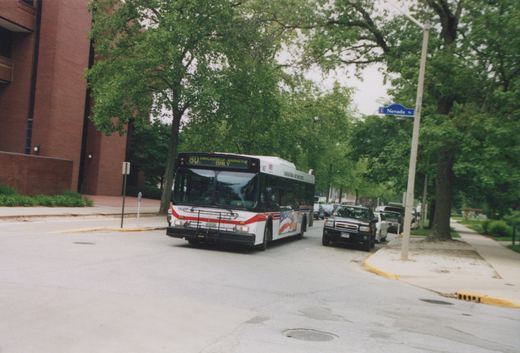 A white and red bus with the number 9667 on the front, labeled Champaign-Urbana Mass Transit District, is making a turn at an intersection with a sign for Nevada Street, surrounded by green trees and parked cars.