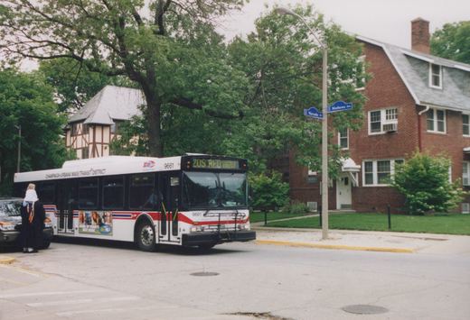 A white and red Champaign-Urbana MTD bus numbered 9661 is stopped on a tree-lined street at an intersection with blue street signs for Nevada St. and Mathews Ave., with a person standing nearby and brick buildings in the background.