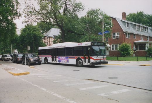 A white, red and blue bus with the number 9667 is turning at a street corner in a residential area, with parked cars and houses in the background.