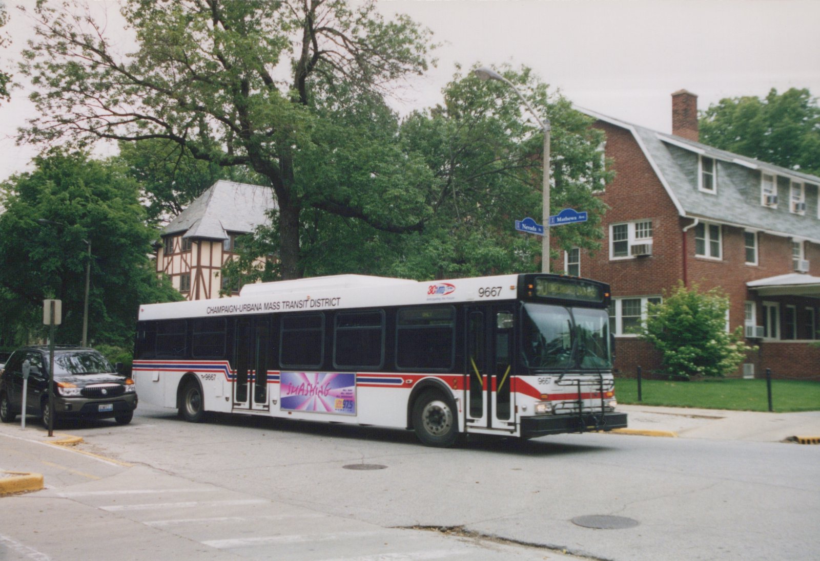 A white and red motor bus with the number 9667 and destination 30 McDonald's is stopped on a street corner near a brick house and street signs for Nevada Street and Mathews Avenue.