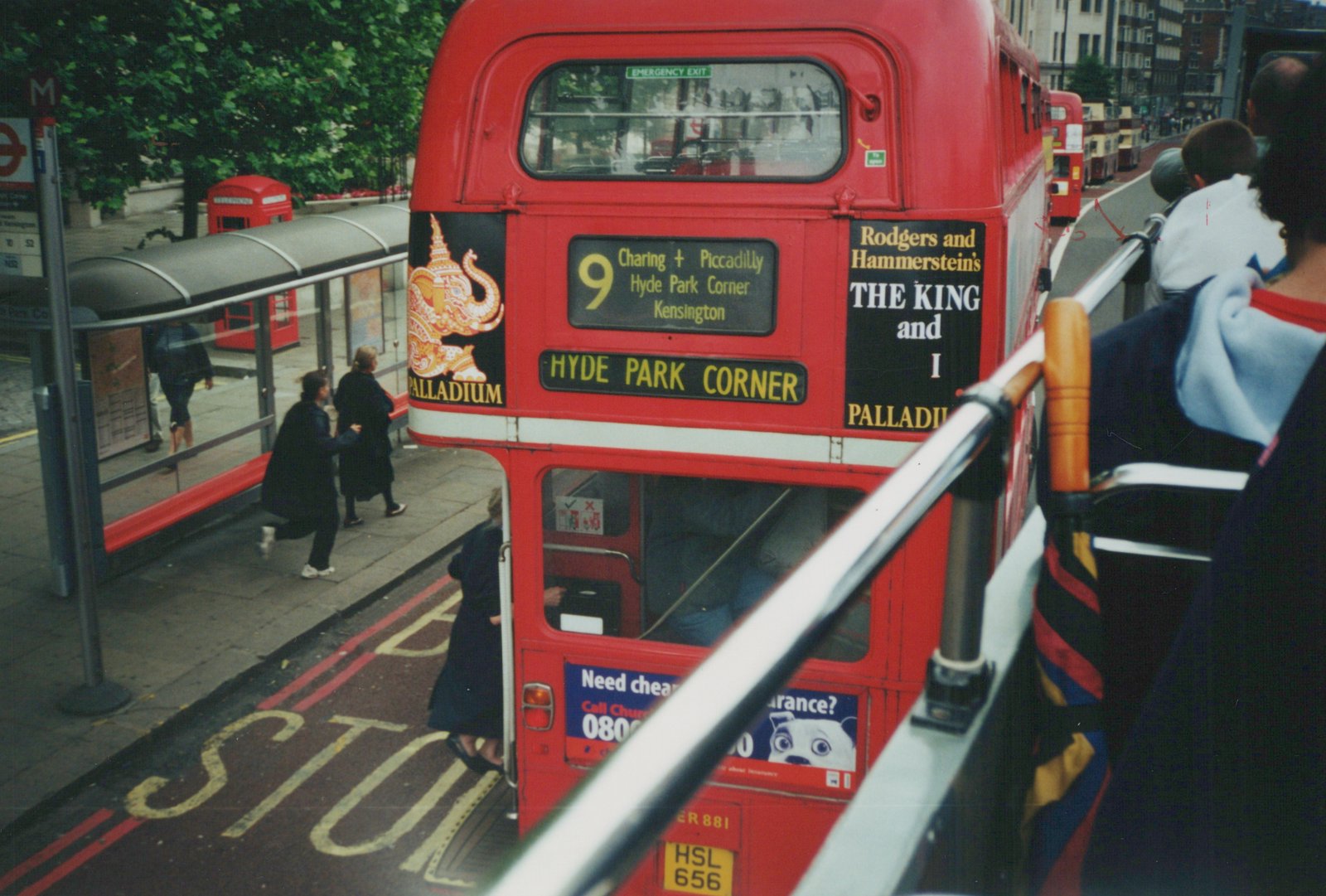 A red double-decker bus is stopped at a bus stop on a city street. People are boarding, and a 'STOP' marking is painted on the road. The bus has a destination of 'Hyde Park Corner' and an ad for 'The King and I'.