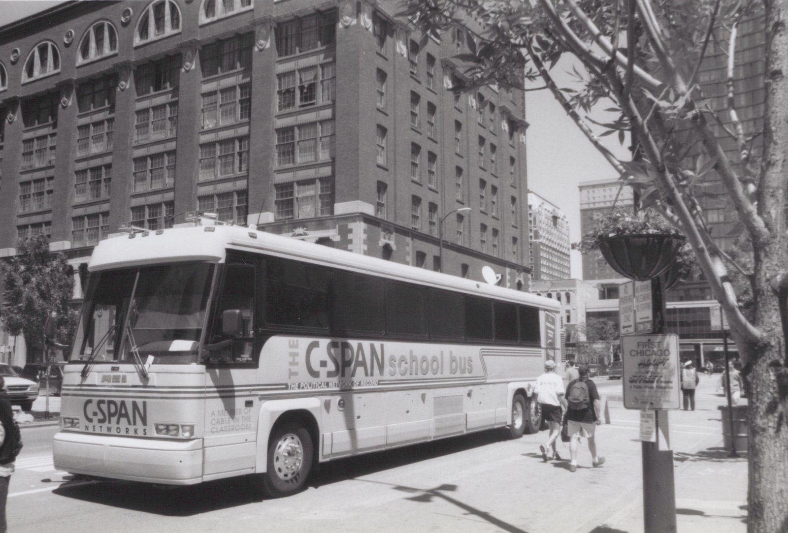 A white C-SPAN school bus is parked on a city street with pedestrians walking nearby and a large brick building in the background.