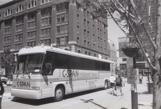 A white C-SPAN school bus is parked on a city street with pedestrians walking nearby and a large brick building in the background.