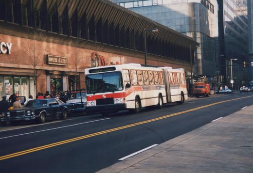 A white and red articulated city bus drives down a street lined with parked cars and modern buildings, with people on the sidewalk.