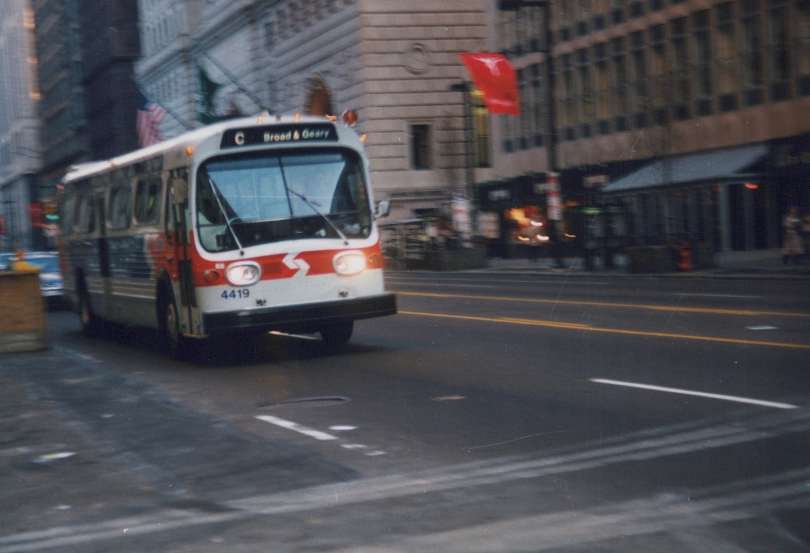 A red and white CTA bus with number 4419 is driving on a city street with buildings and a flag in the background.