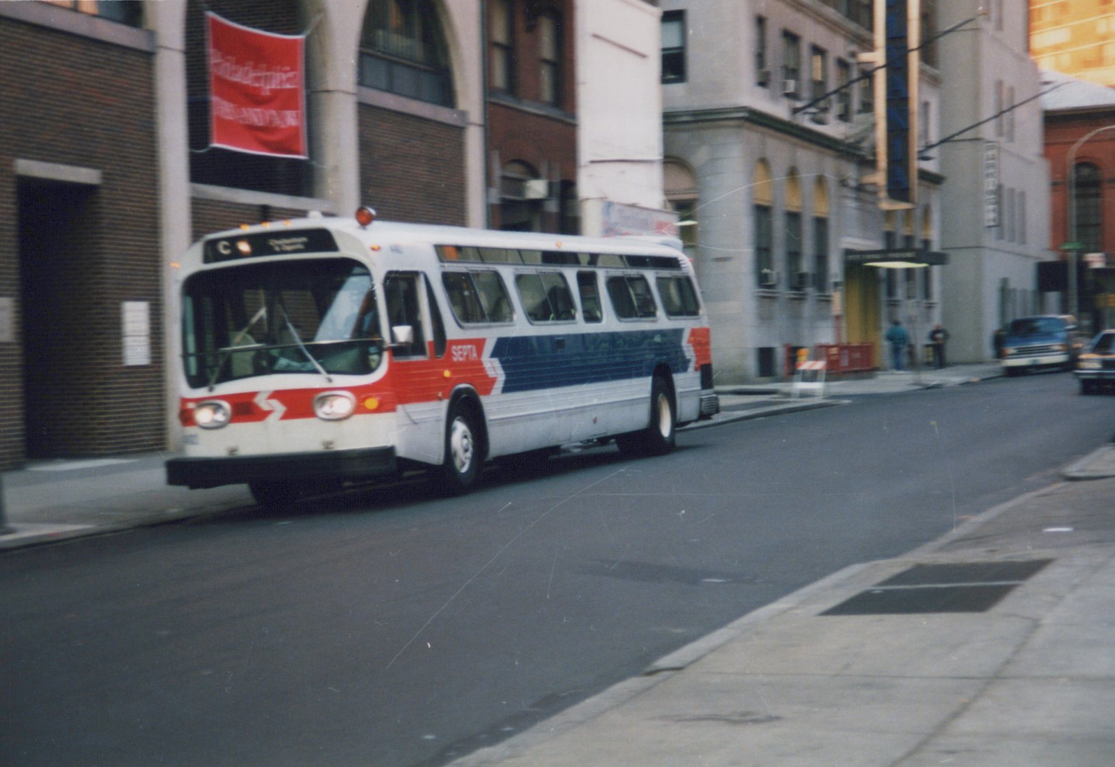 A white, red, and blue SEPTA bus with the number 402 on its front, driving down a city street with buildings and parked cars in the background.