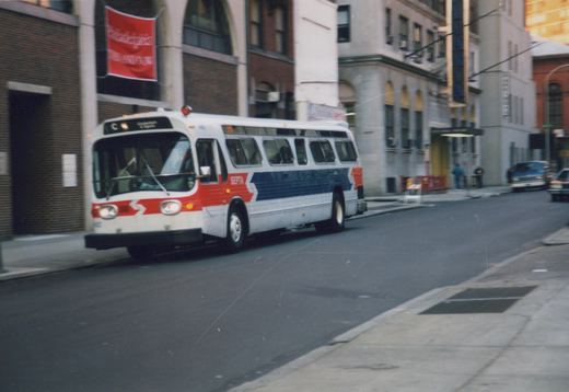 A white, red, and blue SEPTA bus with the number 402 on its front, driving down a city street with buildings and parked cars in the background.