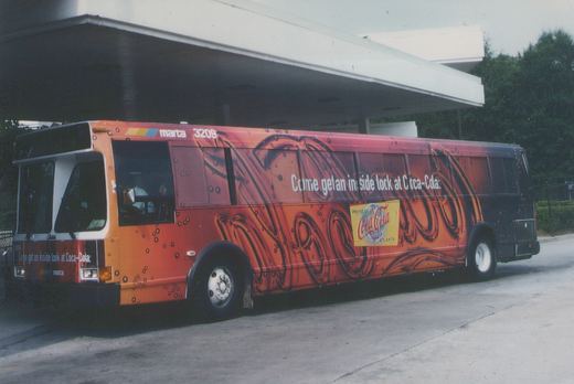 A side view of a MARTA bus with a vibrant orange and black Coca-Cola advertisement, displaying the number 3209, parked under a concrete overpass with trees in the background.