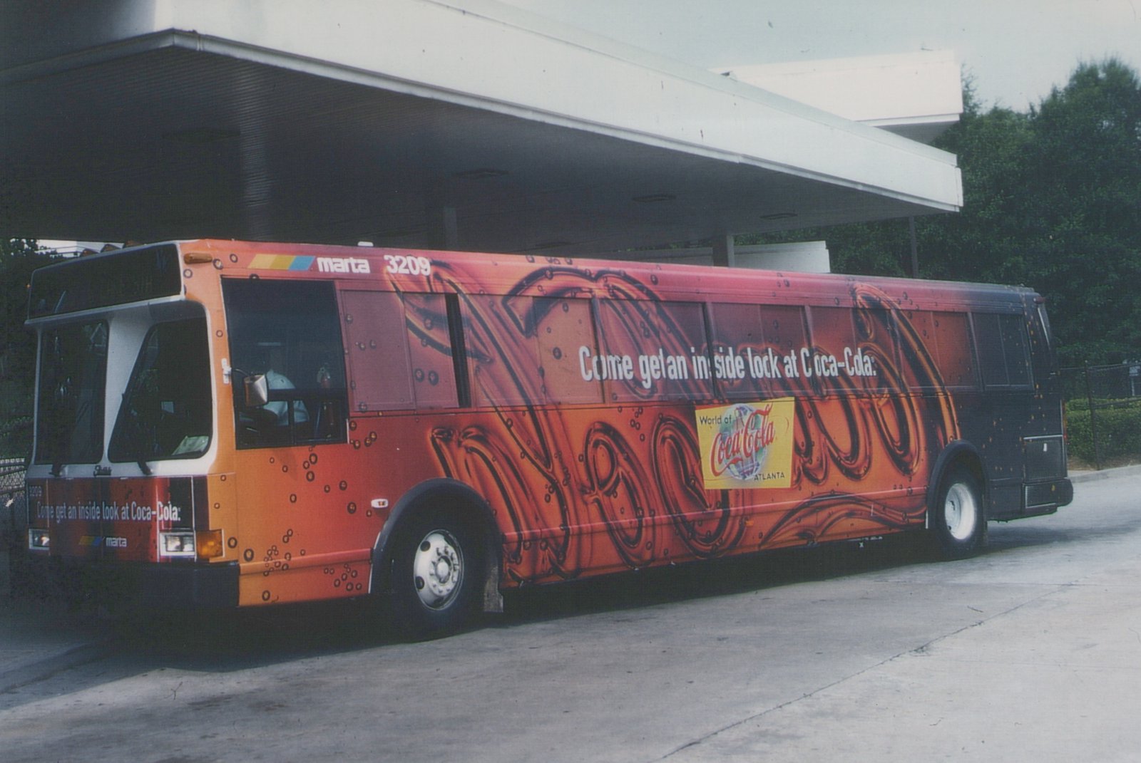 A side view of a MARTA bus with a vibrant orange and black Coca-Cola advertisement, displaying the number 3209, parked under a concrete overpass with trees in the background.