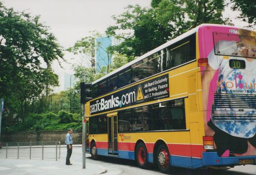 A yellow, red, and blue double-decker Citybus with fleet number 2277 is stopped on a city street under an overcast sky, with a pedestrian nearby and large advertisements on its side and rear.