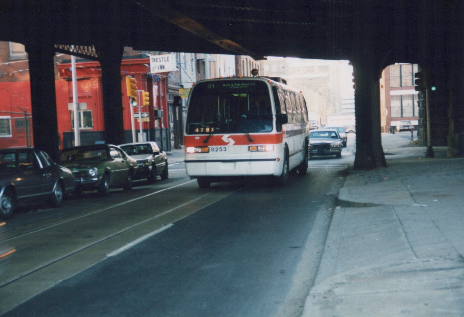 A white and red CTA Flxible Metro bus, number 478X, is driving down a city street under an elevated train structure, with several cars nearby and a red building in the background.