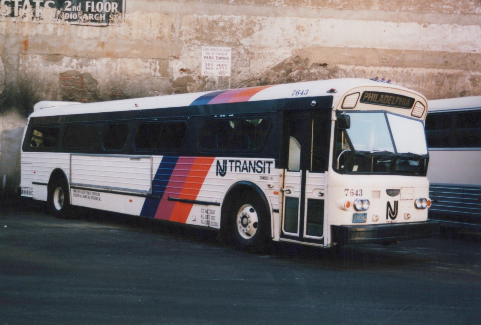 An NJ Transit bus number 7643 with a Philadelphia destination sign, parked on a street with a brick wall and tow-away signs in the background.