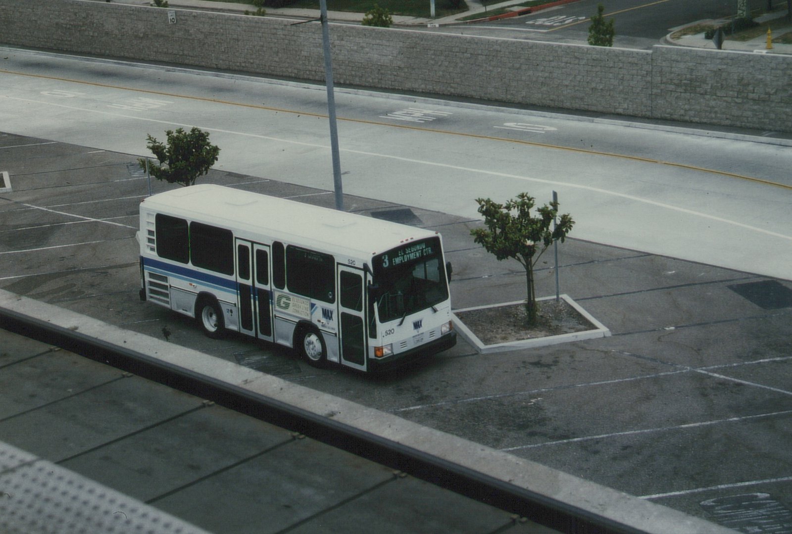A white MAX bus with blue stripes, numbered 520, is parked on a city street beside a parking lot, with its destination sign reading 'El Segundo Employment Center'.