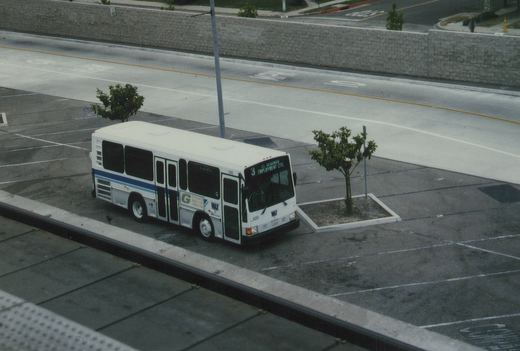 A white MAX bus with blue stripes, numbered 520, is parked on a city street beside a parking lot, with its destination sign reading 'El Segundo Employment Center'.