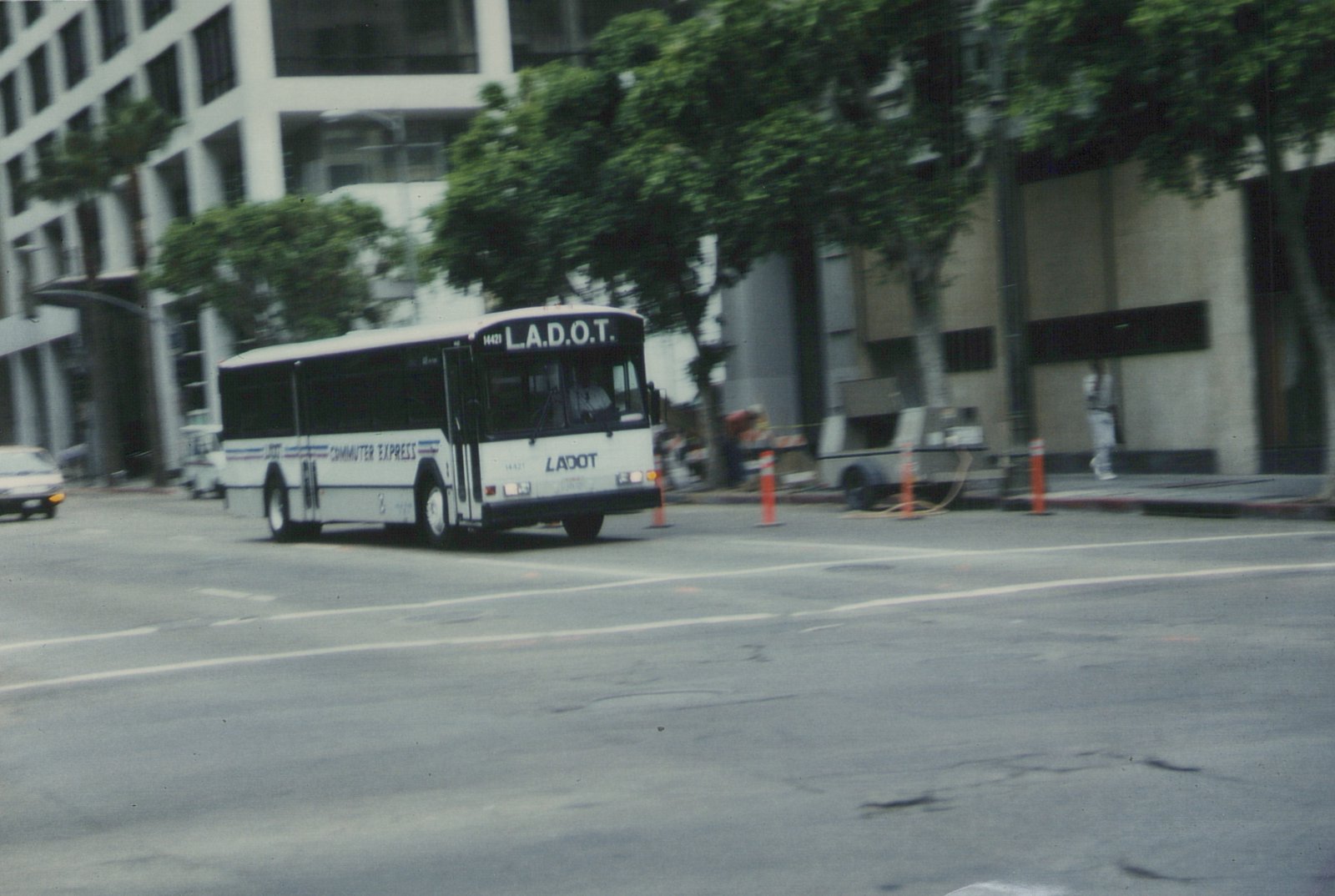 A white LADOT bus with 'COMMUTER EXPRESS' on its side is driving on a city street with buildings and trees in the background.