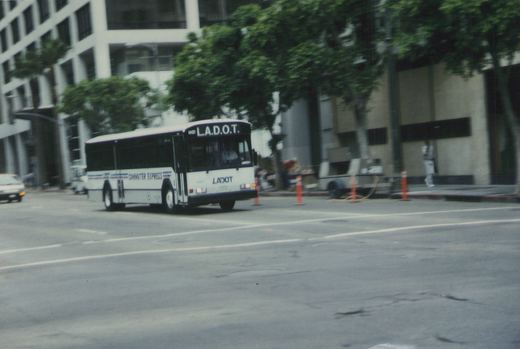 A white LADOT bus with 'COMMUTER EXPRESS' on its side is driving on a city street with buildings and trees in the background.
