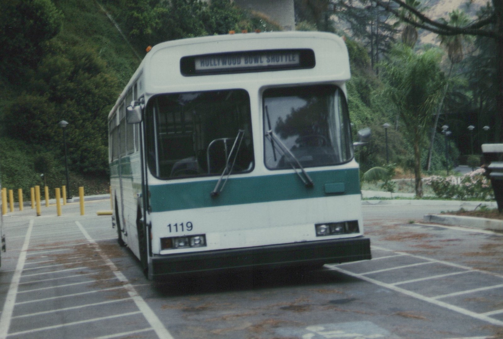 A GM New Look motor bus with the number 1119 and a 'HOLLYWOOD BOWL SHUTTLE' destination sign, parked in a lot with trees and yellow bollards in the background.