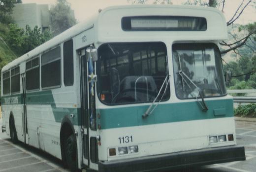 A white and green bus with the number 1131 on its front and side, lettered for Transit Systems, parked on a street with trees and a railing in the background.