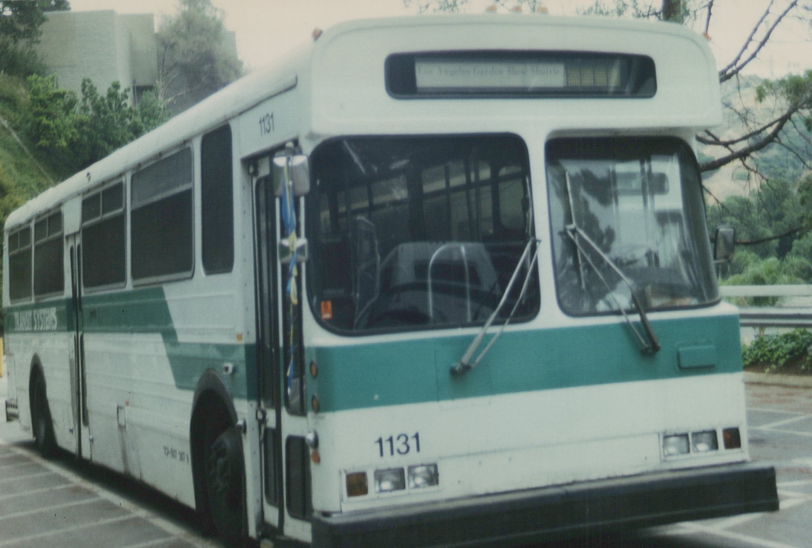 A white and green bus with the number 1131 on its front and side, lettered for Transit Systems, parked on a street with trees and a railing in the background.