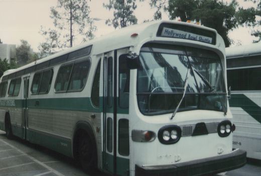 A white and green bus with 'Hollywood Bowl Shuttle' on the front sign, marked 'Transit Systems' on the side, parked on a street with trees in the background.
