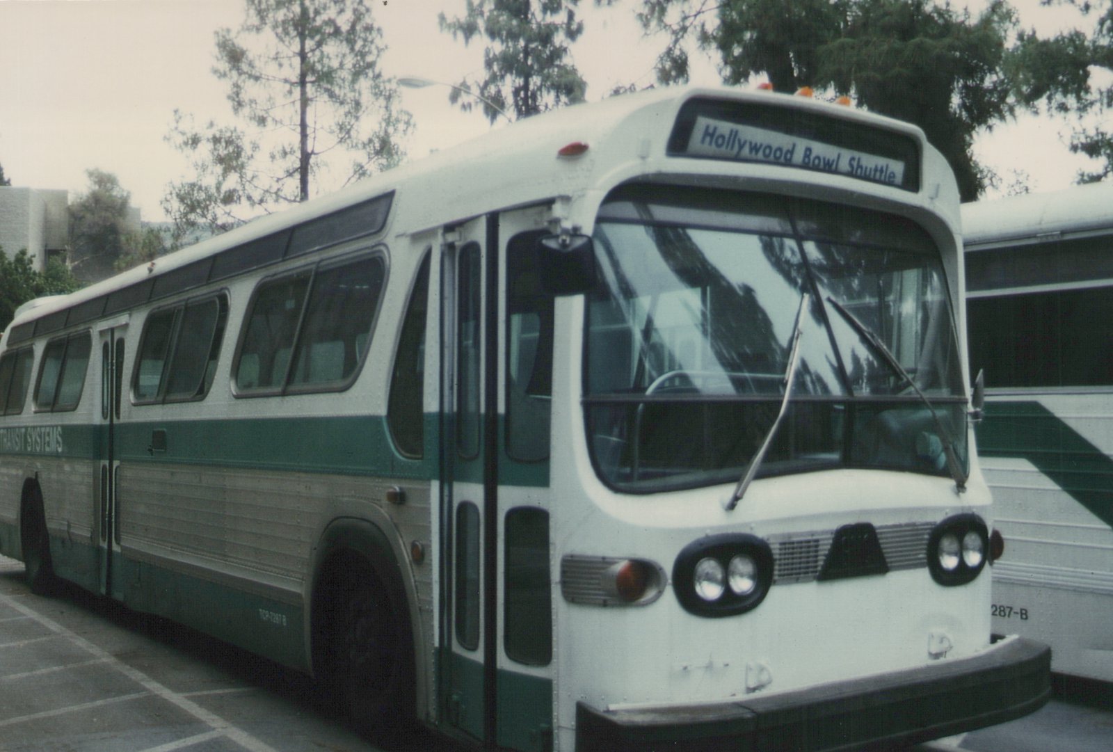 A white and green bus with 'Hollywood Bowl Shuttle' on the front sign, marked 'Transit Systems' on the side, parked on a street with trees in the background.