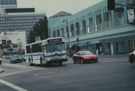 A white Foothill Transit bus with blue stripes is driving on a city street, with a red car and a building with 'GLACIAL' on its sign in the background.