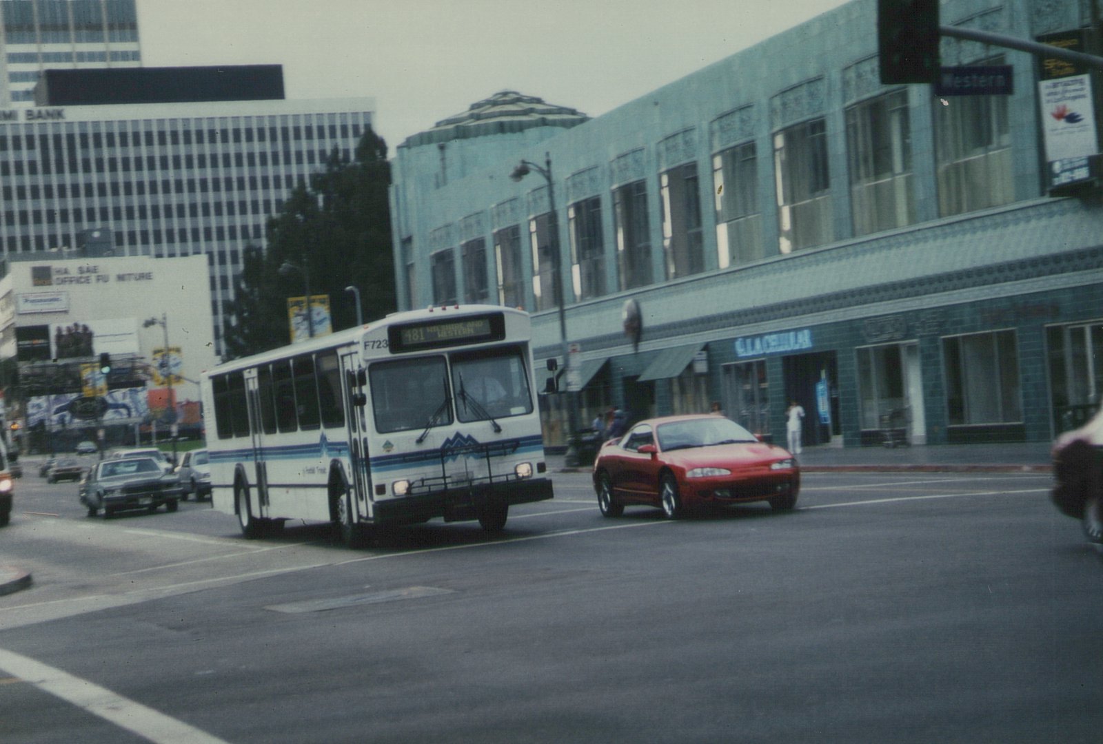 A white Foothill Transit bus with blue stripes is driving on a city street, with a red car and a building with 'GLACIAL' on its sign in the background.